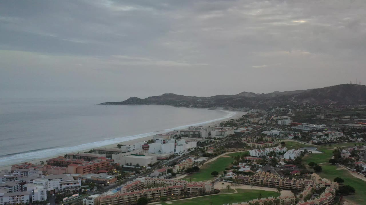 Elevated Los Cabos coastal view shows a pale beach curve beside the gray ocean, compact buildings and roads inland, green fairways, distant hills, and no visible people.