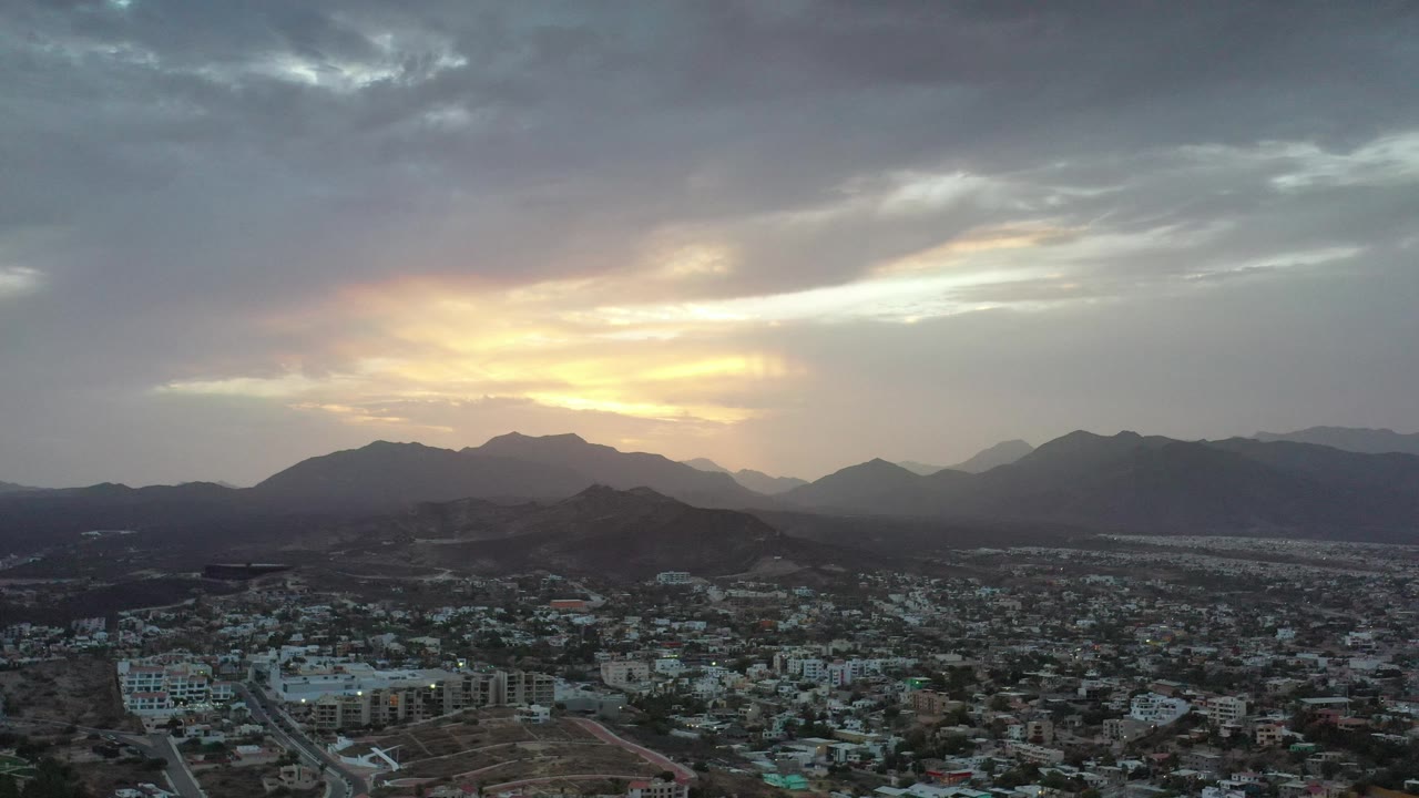 Los Cabos aerial frame showing low-rise urban development across the lower half, dark hills and mountains in the distance, and a bright break in the cloud cover with no visible people.