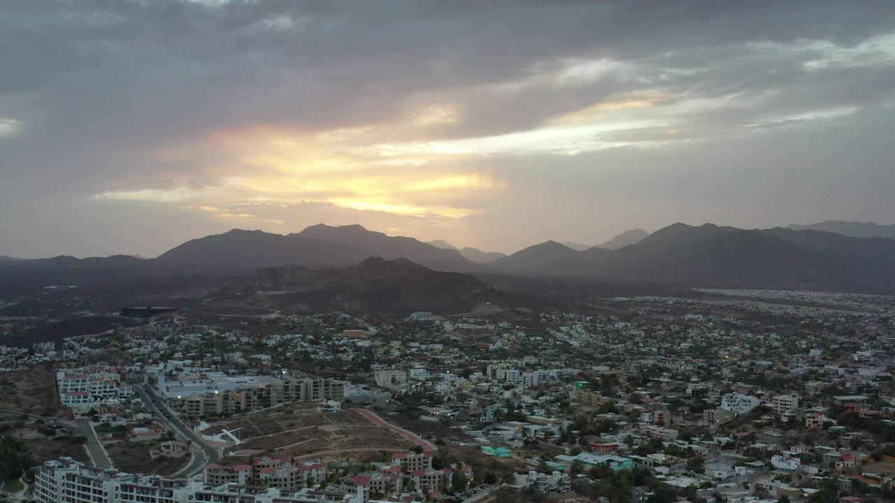 Wide aerial view over Los Cabos, Mexico, with rooftops and roads below dark mountain ridges, golden light in the clouds, and no visible people.