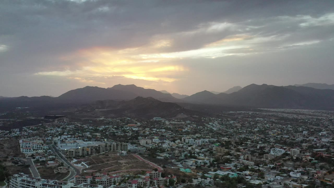 High view of Los Cabos buildings and hills under a cloudy sunset.