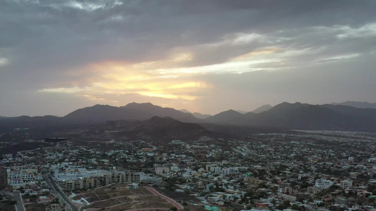 Second aerial frame of Los Cabos with the city spread across the lower frame, mountain silhouettes on the horizon, brighter gold cloud breaks, and no visible people.