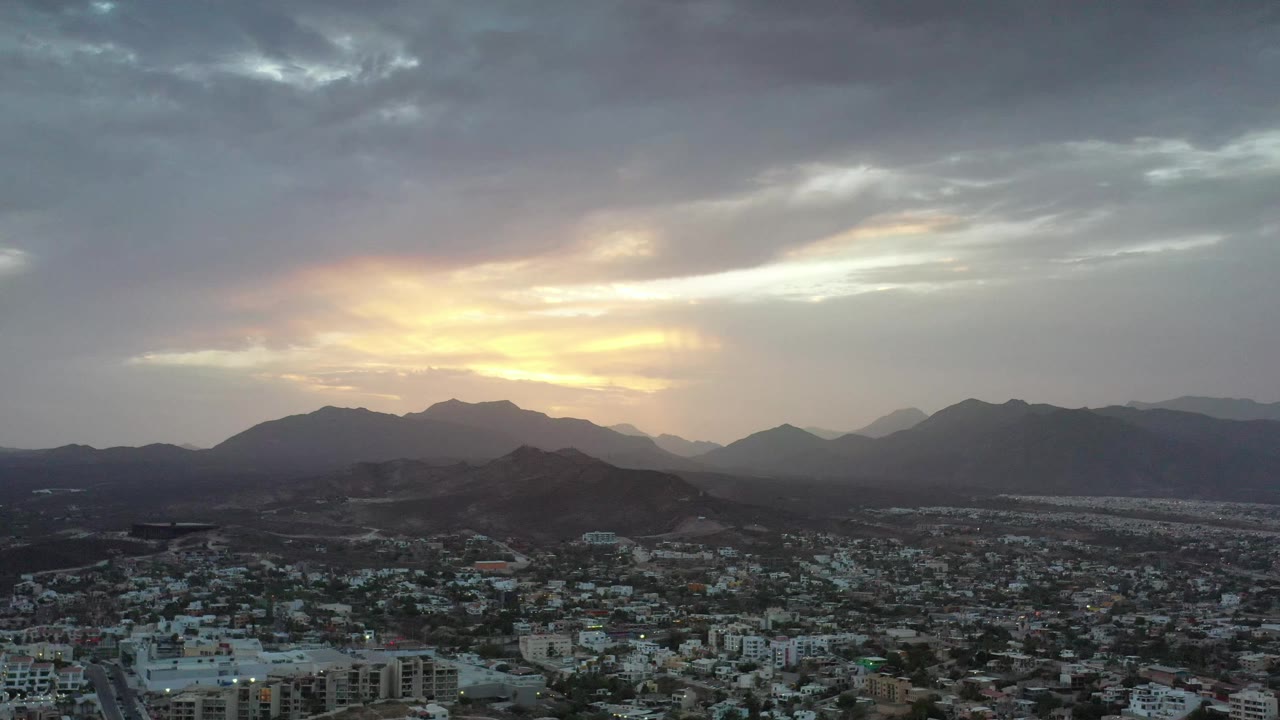 A similar aerial moment over Los Cabos shows the city grid and homes in the lower frame, dark hills across the middle distance, glowing sunset clouds, and no visible people.