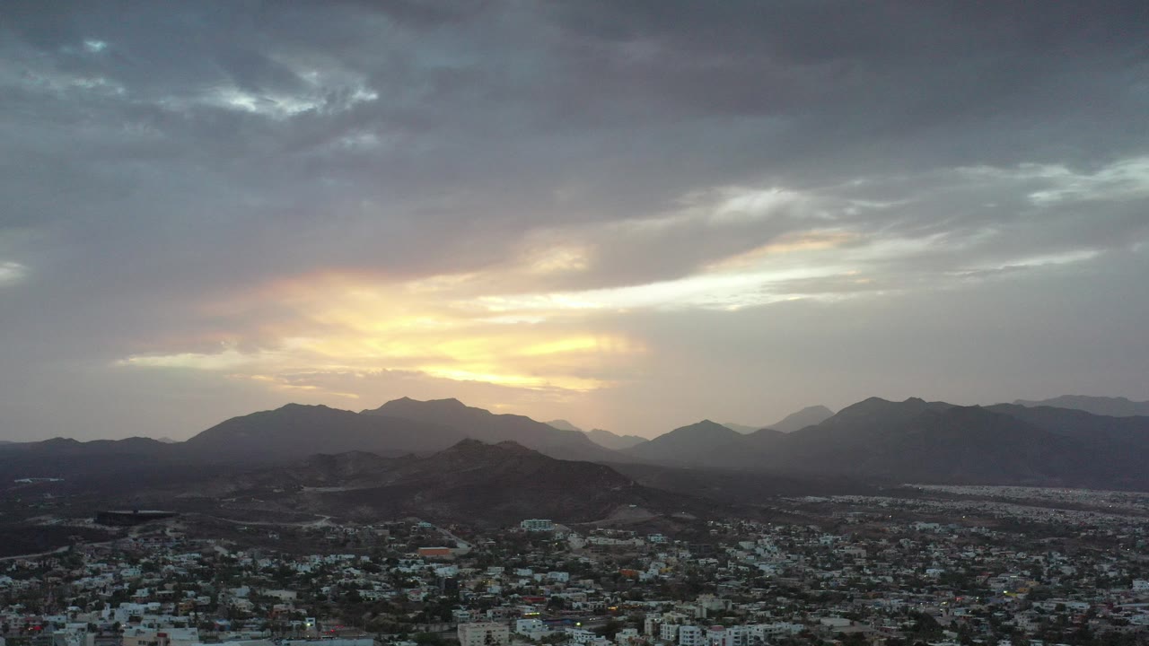 Wide elevated view over Los Cabos, Mexico, with city buildings in the foreground, dark mountain ridges beyond, yellow sunset light breaking through gray clouds, and no visible people.