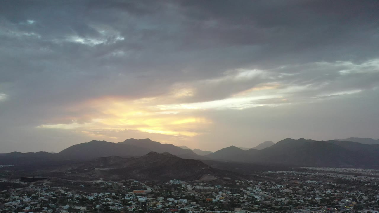 A wide Los Cabos aerial view shows dense low buildings below layered mountains, with brighter yellow sunset light spreading through gray clouds and no visible people.