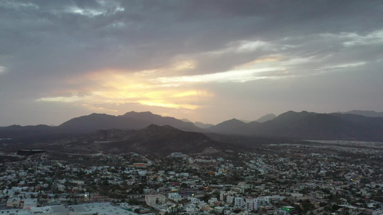 Aerial view of a town below dark hills and a cloudy sunset sky.