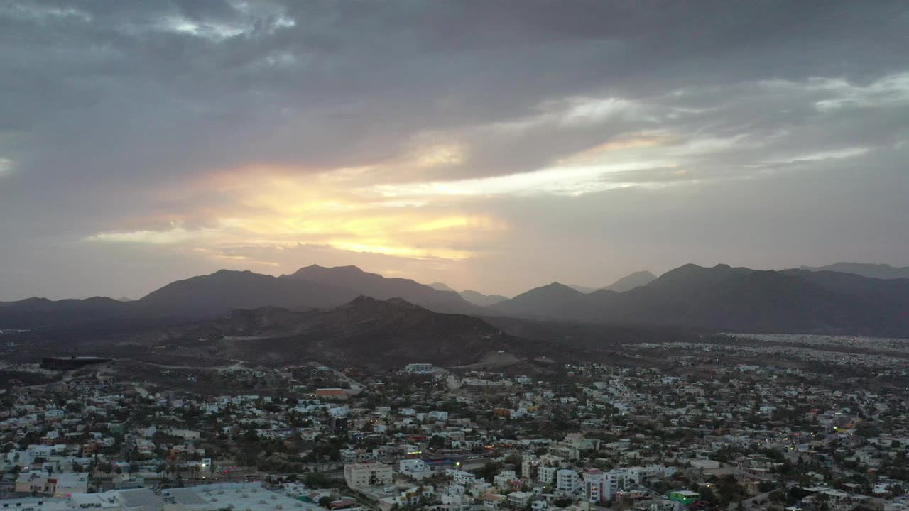 Early frame over Los Cabos, Mexico, with dense low buildings in the foreground, shadowed hills in the middle distance, and a bright sunset opening behind clouds, no visible people.