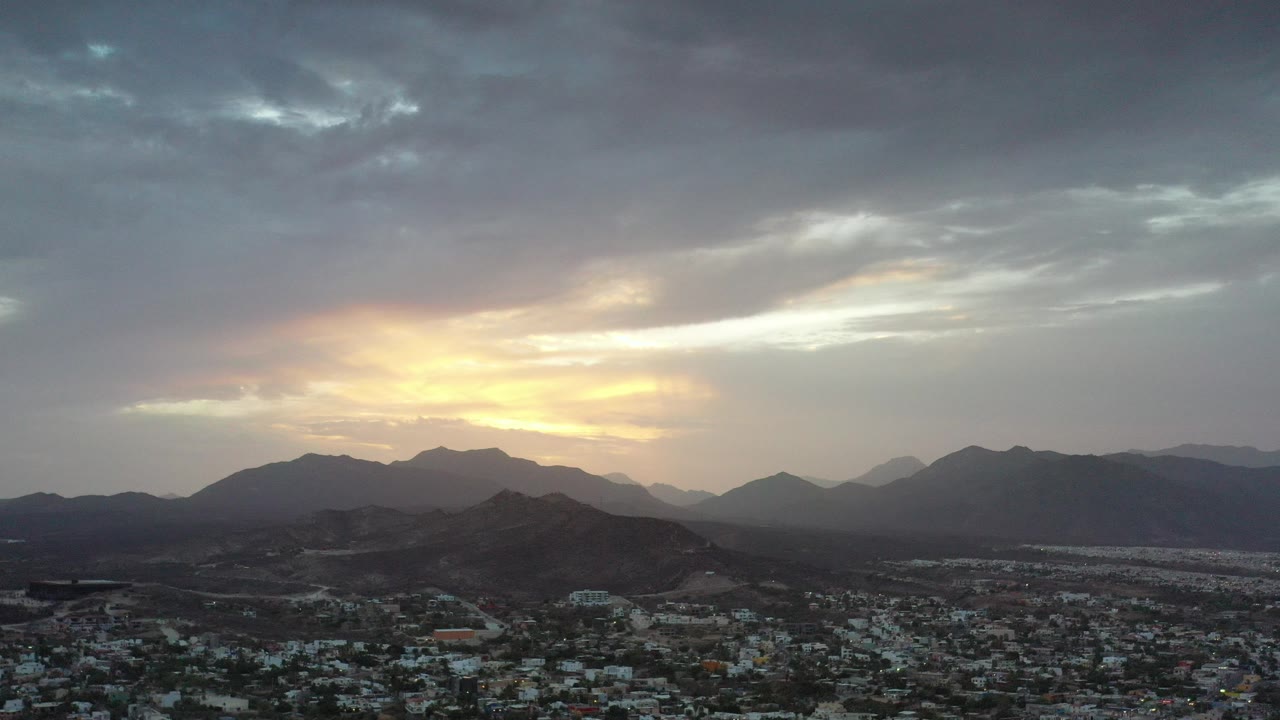 Los Cabos sunset view with a low city foreground, dark mountain ridges, a yellow horizon glow, layered gray clouds, and no visible people.