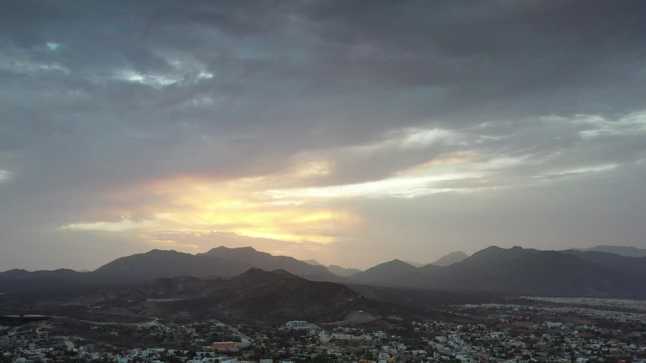Los Cabos, Mexico sunset with a bright yellow opening in layered gray clouds above dark mountain ridges and a town below, no visible people.