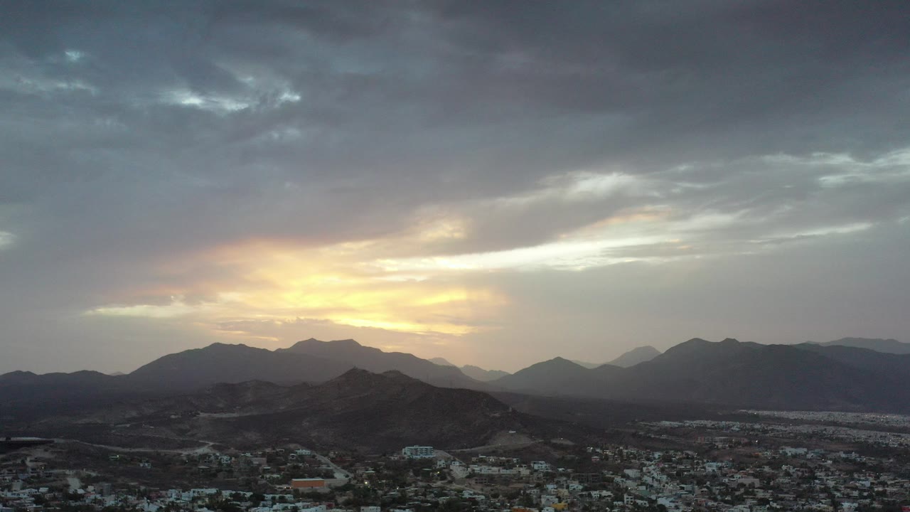 Opening aerial frame over Los Cabos, Mexico, with a town at the bottom edge, dark mountain ridges beyond, warm sunset light through gray clouds, and no visible people.