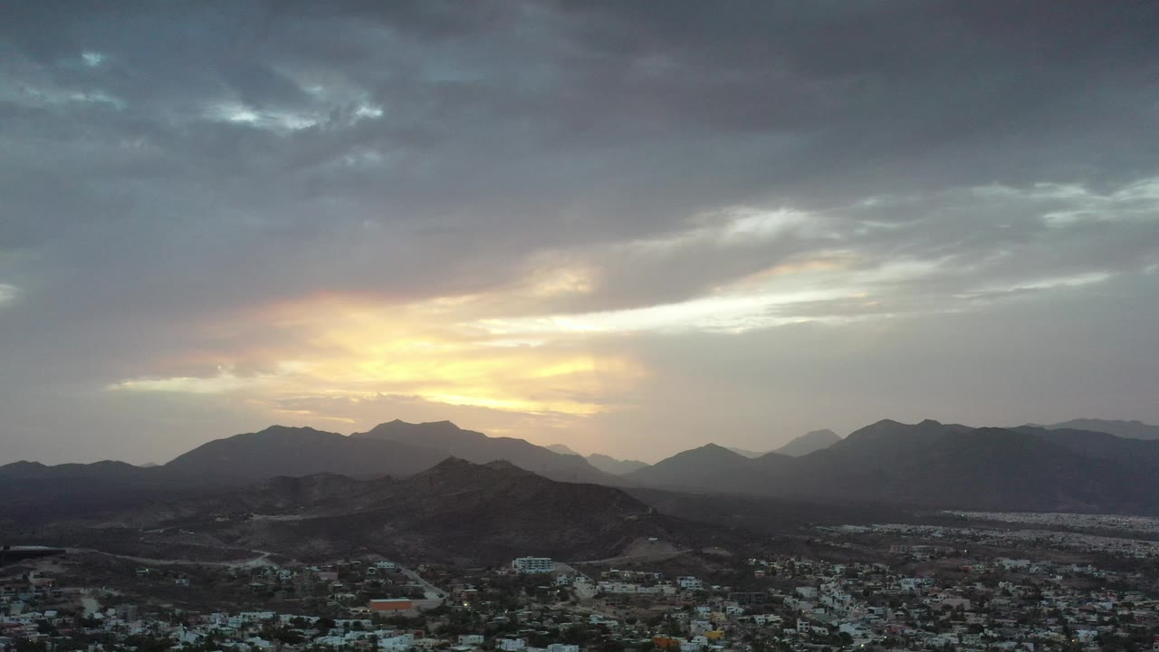 Wide aerial landscape with town, hills, mountains, and glowing clouds.