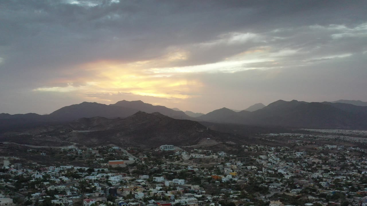 Wide aerial view of a city below dark mountains at sunset.