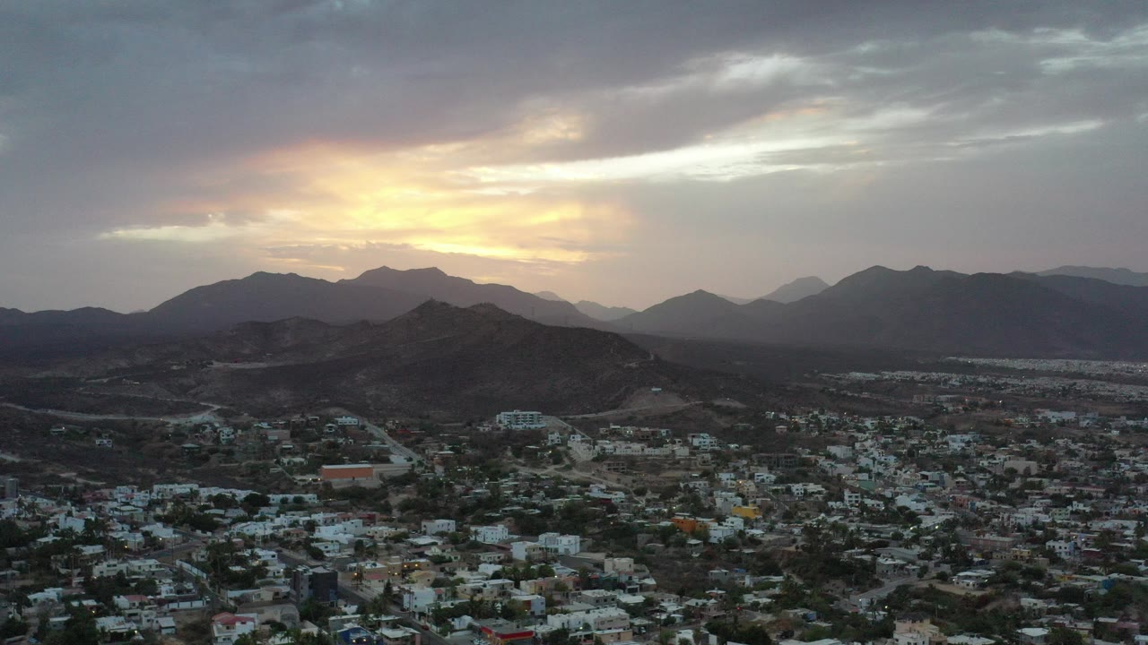 Los Cabos city below mountains at sunset under gray clouds.