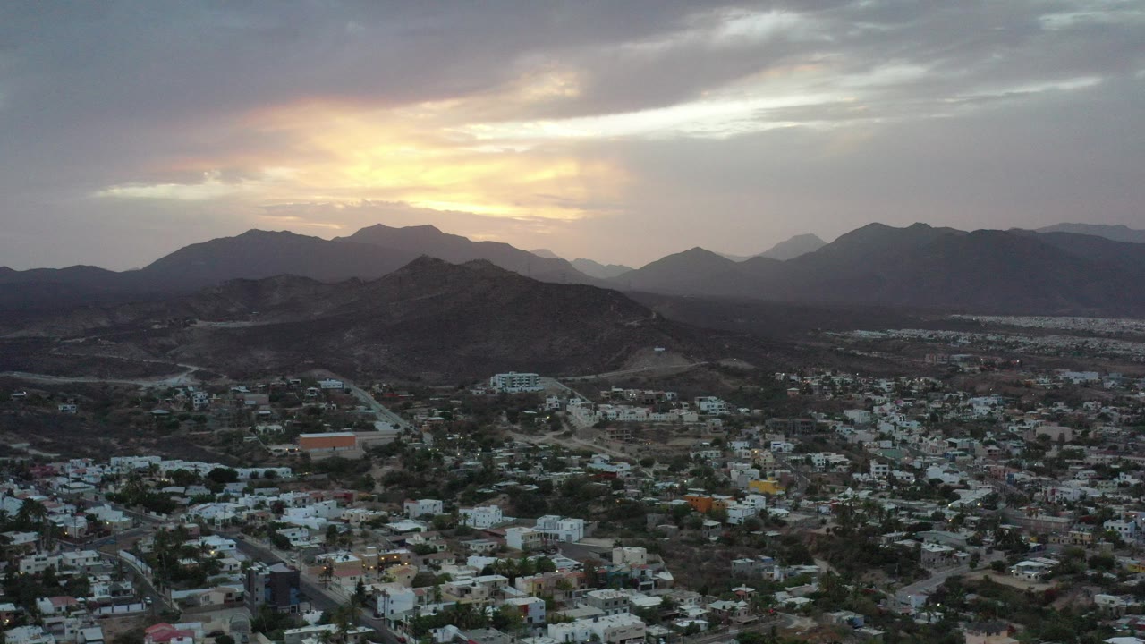 Los Cabos, Mexico, appears from an elevated wide view with low buildings in the foreground, dark hills beyond, yellow sunset light under gray clouds, and no visible people.