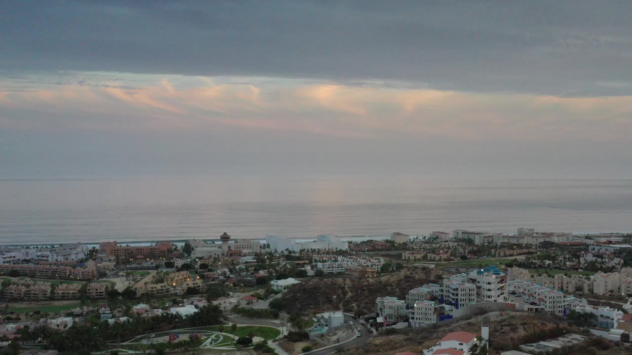 Opening aerial frame over Los Cabos, Mexico resort buildings and shoreline under a heavy gray morning sky, with no visible people.