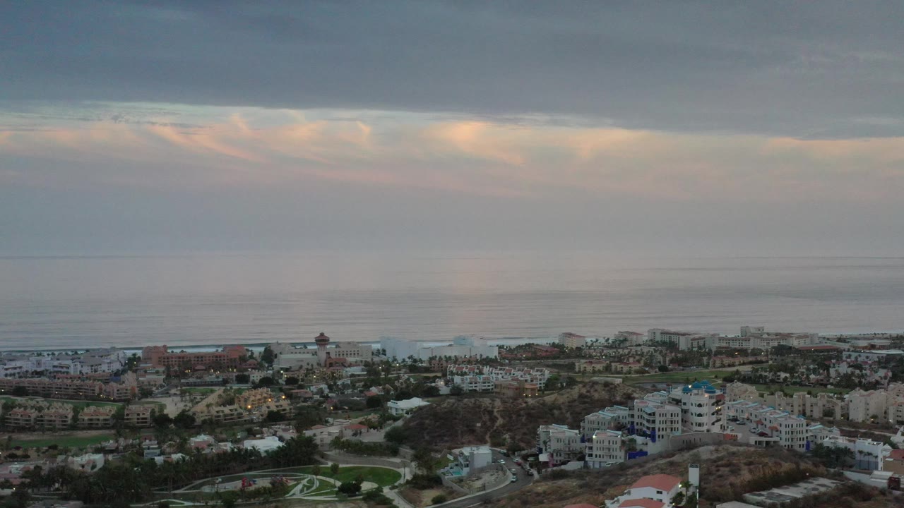 Aerial Los Cabos, Mexico frame with resort blocks, green grounds, a pale shoreline, and soft peach light beneath gray clouds, with no visible people.
