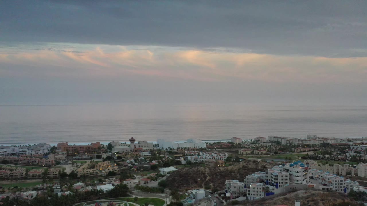 Aerial frame in Los Cabos, Mexico, showing oceanfront residential buildings, roads, and a broad gray cloud deck with no visible people.