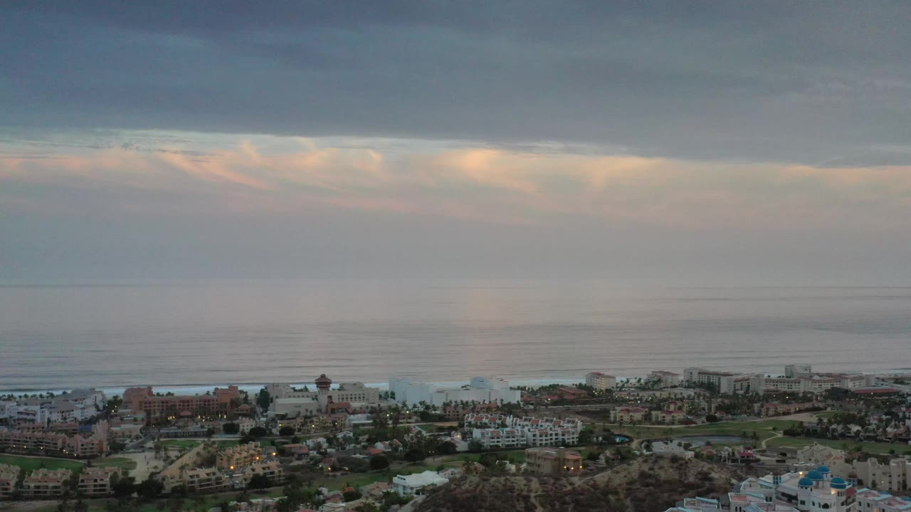 Los Cabos coastline seen from above, with muted sunlight under a dark cloud layer, low surf near shore, and no visible people.