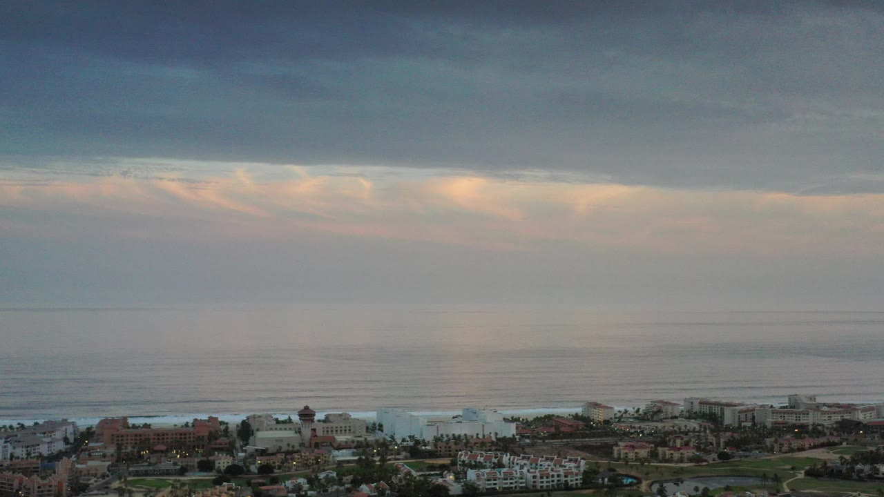 Los Cabos aerial view with city buildings along the bottom edge, a broad gray sea, layered clouds, and no visible people.