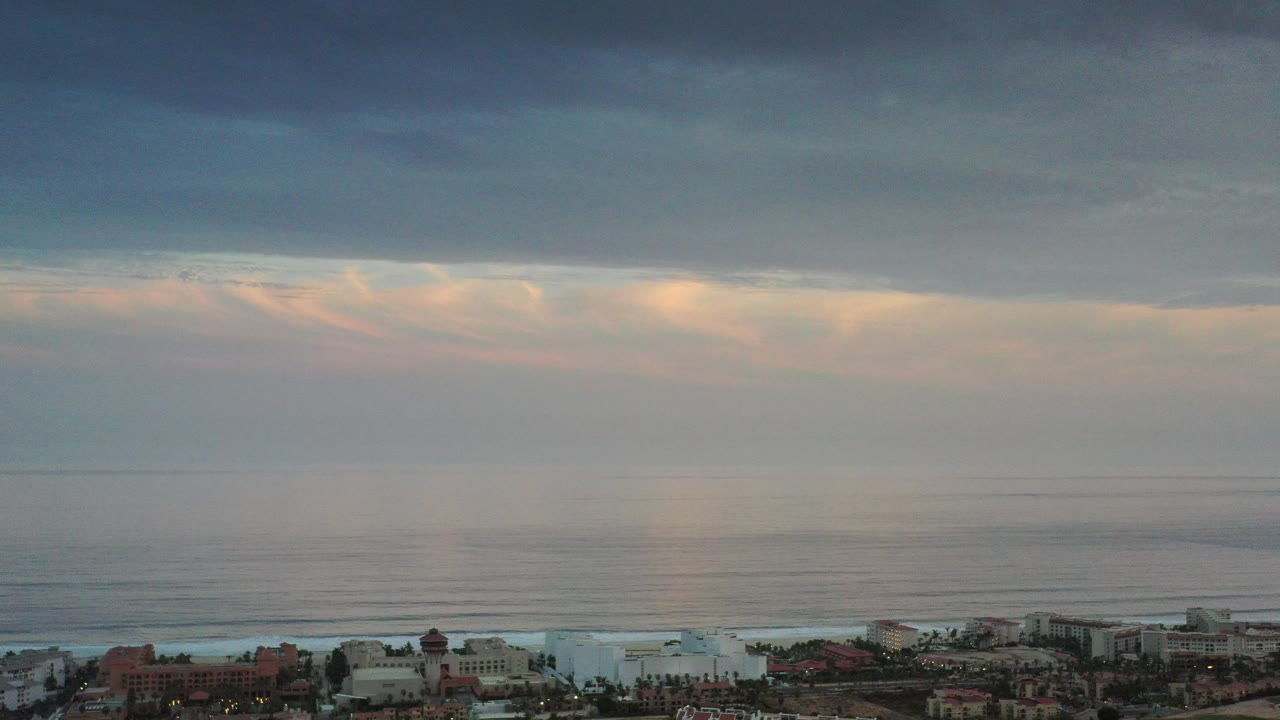 Los Cabos, Mexico dusk view with beachfront buildings along the bottom edge, smooth ocean bands, warm light near the horizon, dark cloud cover overhead, and no visible people.