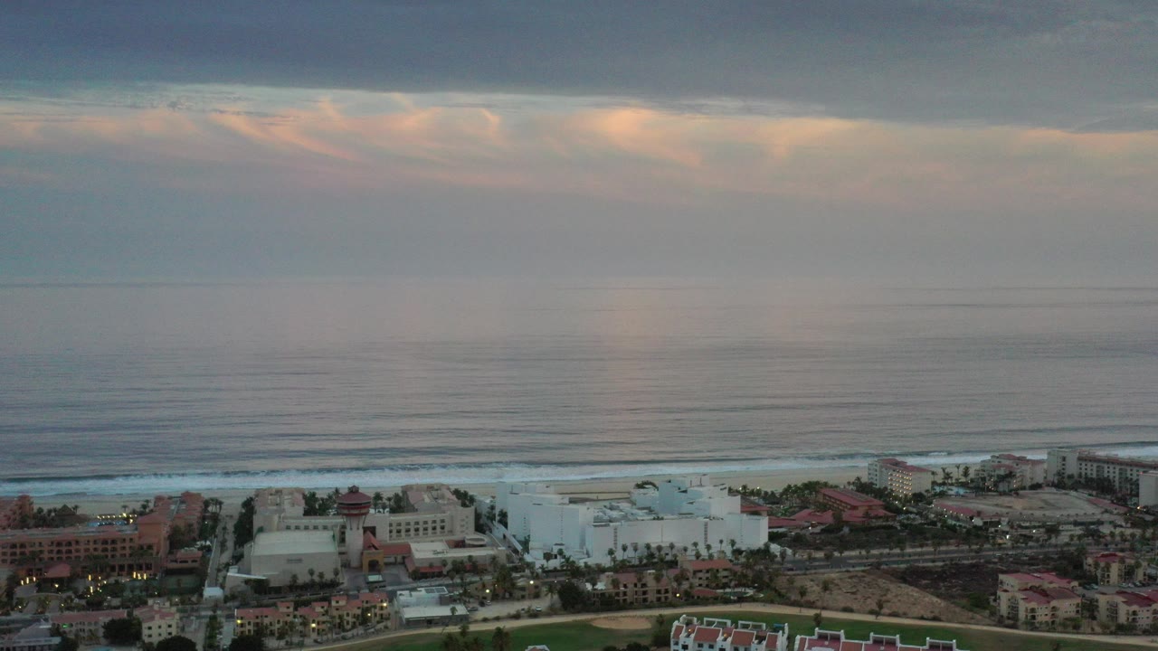 Aerial view of ocean surf and beachfront buildings under layered clouds.