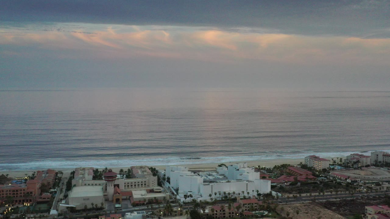 Aerial view of coastal buildings beside a beach and calm ocean under clouds.
