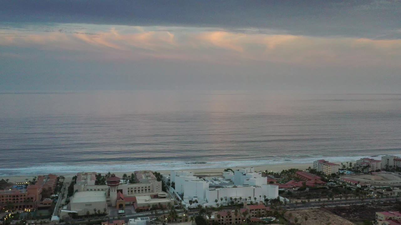 Los Cabos, Mexico aerial view with white beachfront buildings, red roof sections, a thin surf line, wide ocean surface, layered clouds, and no visible people.