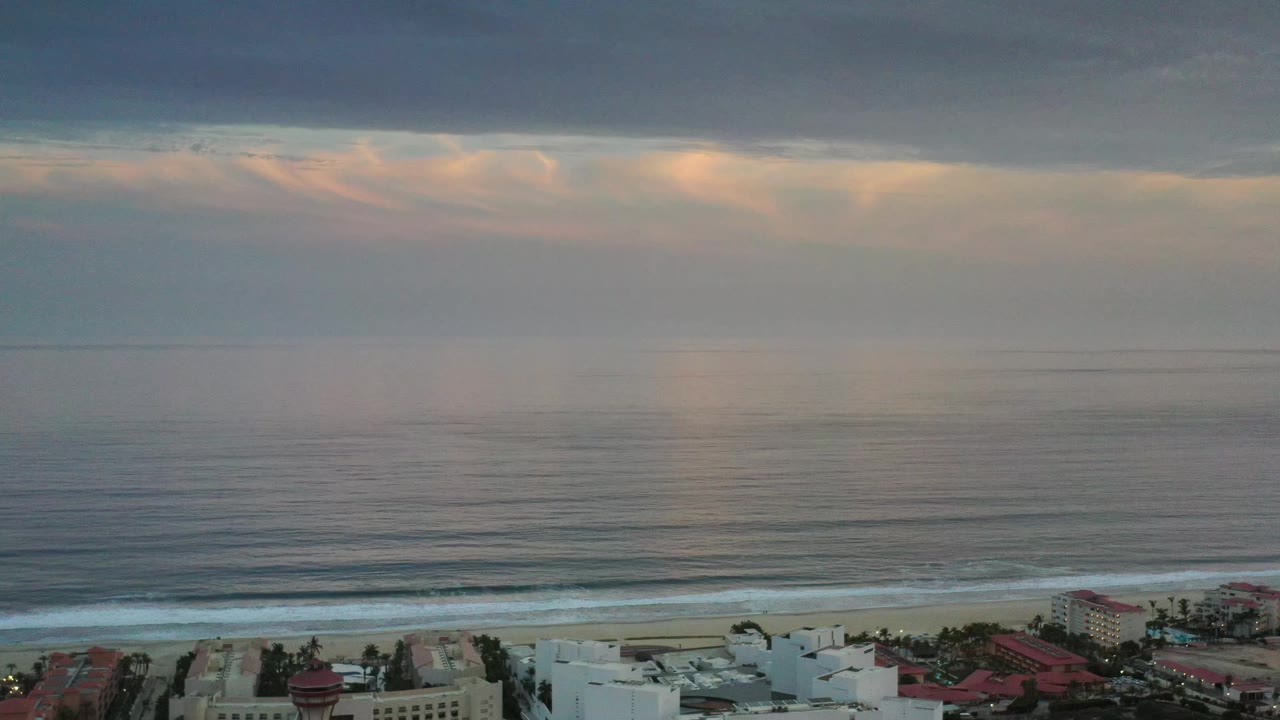 A Los Cabos shoreline view shows muted open water beneath pink and peach haze, a dark cloud band across the top, surf rolling toward the sand, and no visible people.