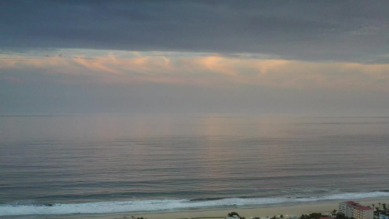 Los Cabos coastline seen from above with rippled blue-gray water, pale surf near the beach, a warm break in the cloud layer, and no visible people.