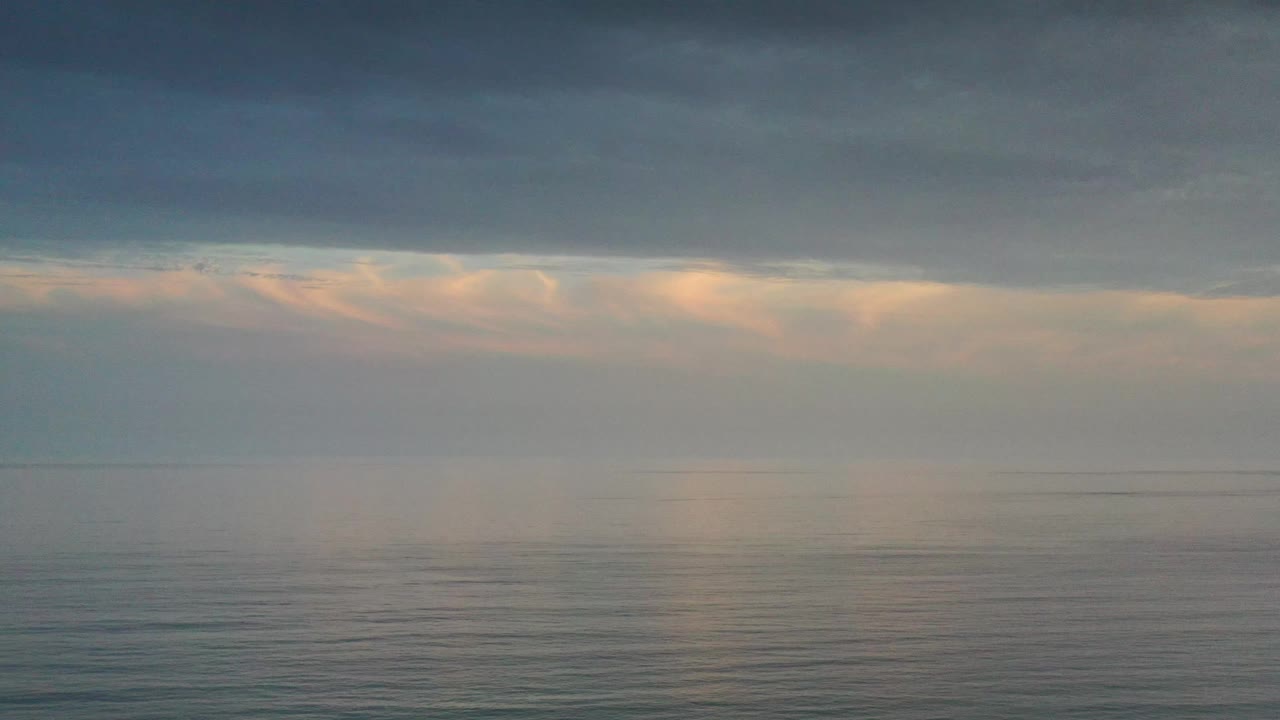 A calm Los Cabos ocean surface reflects muted peach light below a broad cloud shelf, with the horizon running level and no visible people.