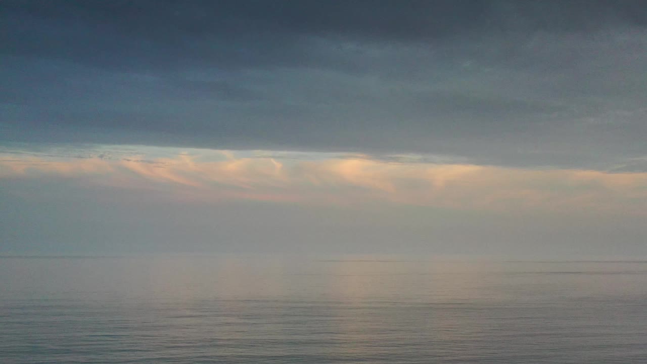 Los Cabos ocean sunrise view one second in, showing still blue-gray water, a low horizon, orange cloud glow, and no visible people.