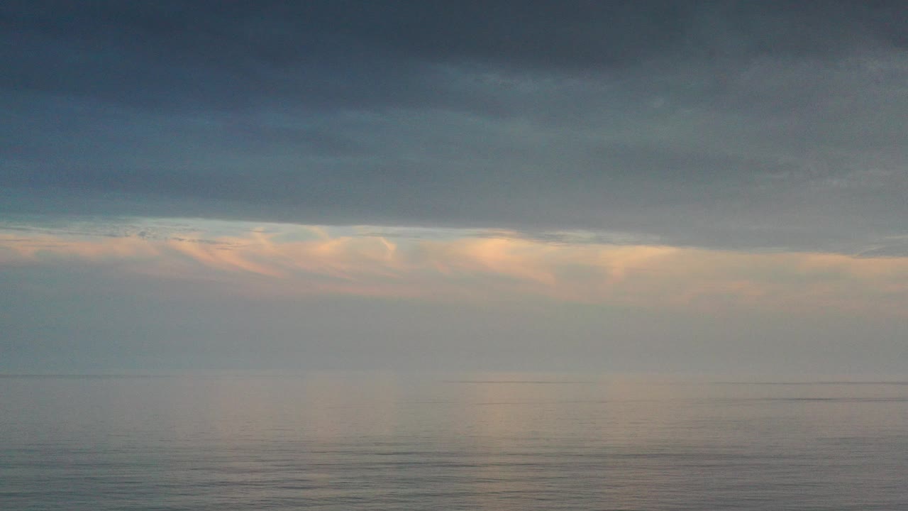 Opening view of the Los Cabos ocean horizon under a dark gray cloud band, with warm light breaking through low clouds and no visible people.