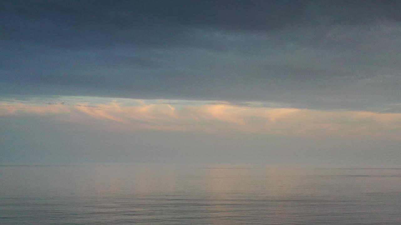 A wide Los Cabos seascape shows a heavy cloud ceiling over calm water, with a peach band of light near the horizon and no visible people.