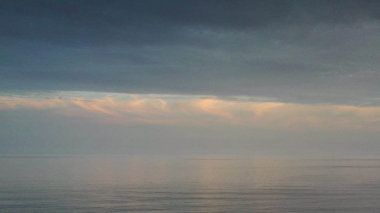 Opening frame of a calm Los Cabos ocean horizon under a broad blue-gray cloud layer, with peach sunset light low in the sky and no visible people.