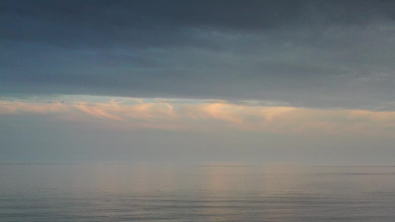 A Los Cabos ocean horizon remains centered as warm gold and peach light spreads through lower clouds below a darker overcast band, with no visible people.