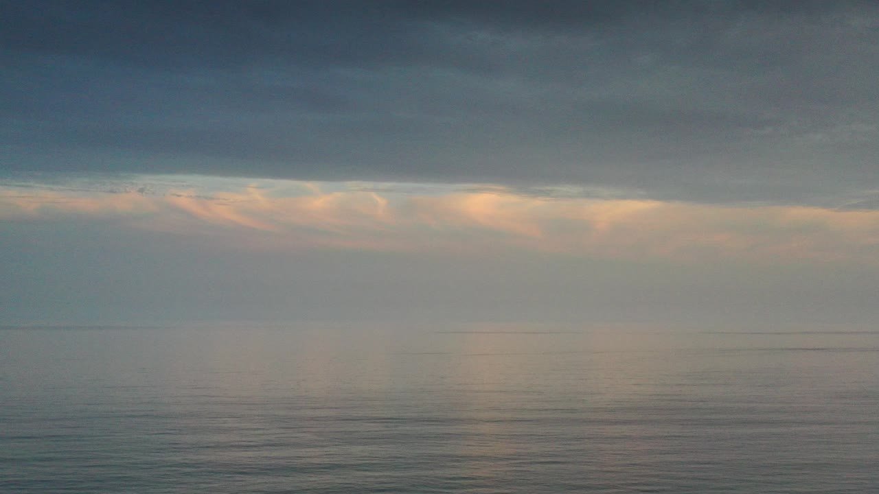 Opening frame of the Los Cabos ocean horizon with smooth blue-gray water, a dark cloud layer overhead, peach light on distant clouds, and no visible people.