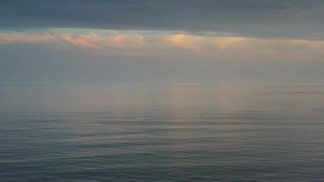 Wide Los Cabos ocean view at the start of the clip, with muted blue water, a hazy horizon, peach light in the clouds, and no visible people.