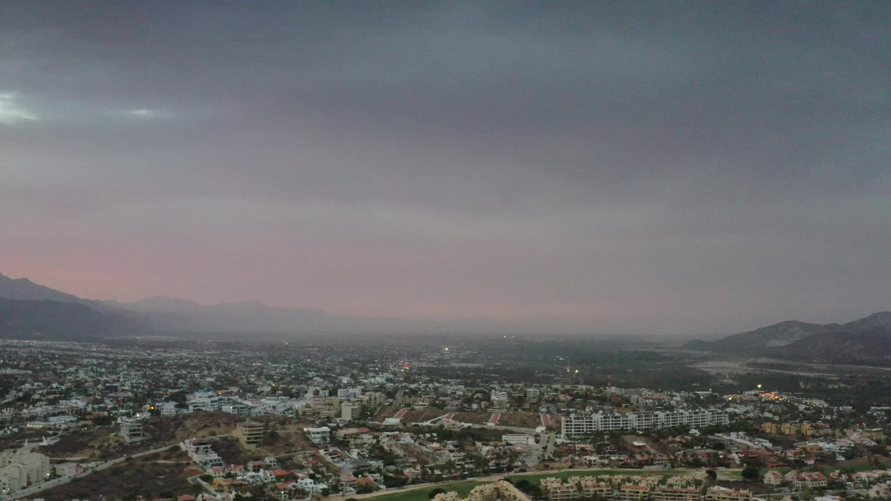 Aerial view over Los Cabos, Mexico, with the city filling the lower frame, mountains framing the valley, soft pink light at the horizon, and no visible people.
