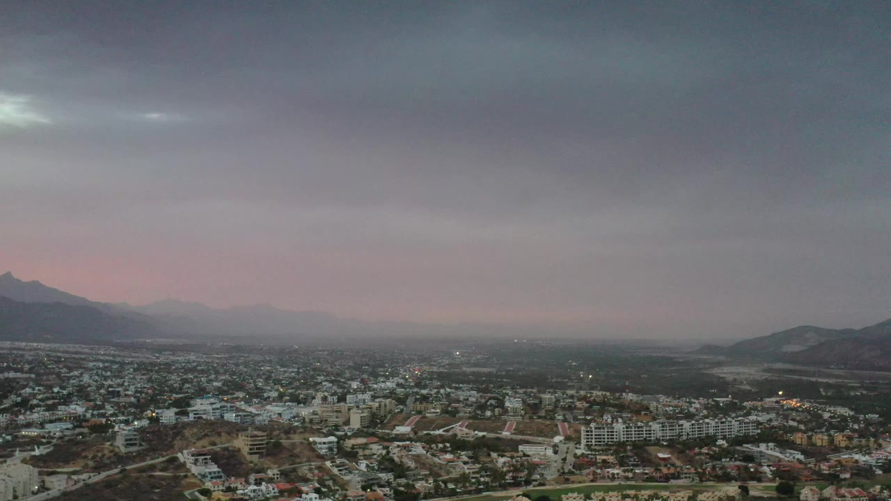 Early frame over Los Cabos, Mexico, showing clustered buildings and roads beneath a broad gray sky, with mountains silhouetted at the left and right and no visible people.