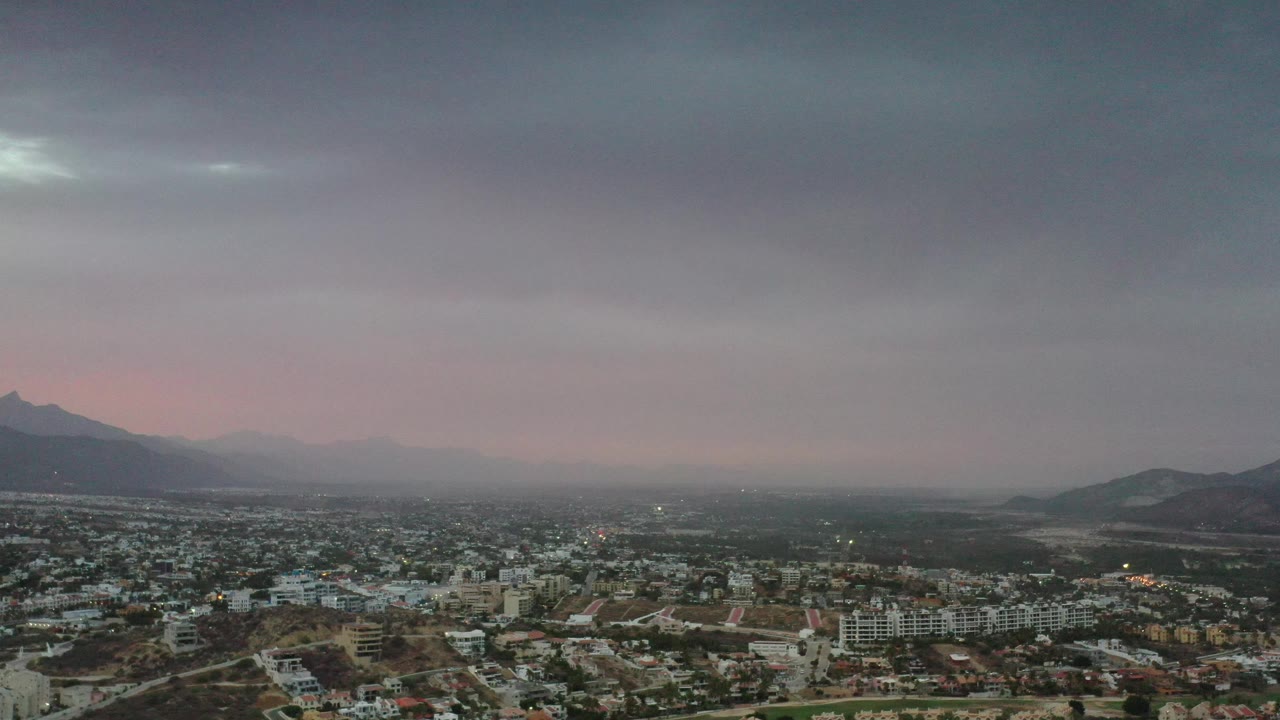 Los Cabos, Mexico, appears in a wide elevated composition, with residential and commercial-looking buildings below, dark ridgelines at the edges, soft pink light near the horizon, and no visible people.