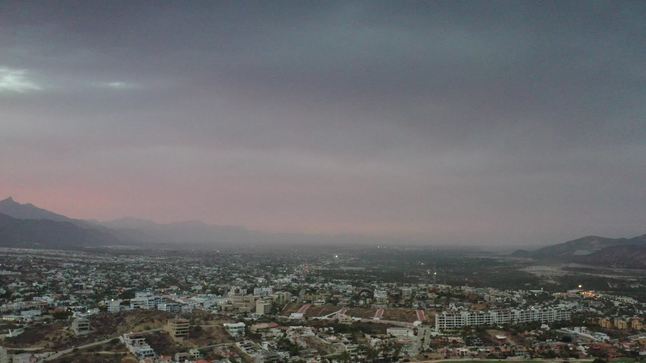 Opening frame of Los Cabos from above, with city buildings in the foreground, hazy mountains and water in the distance, and no visible people.