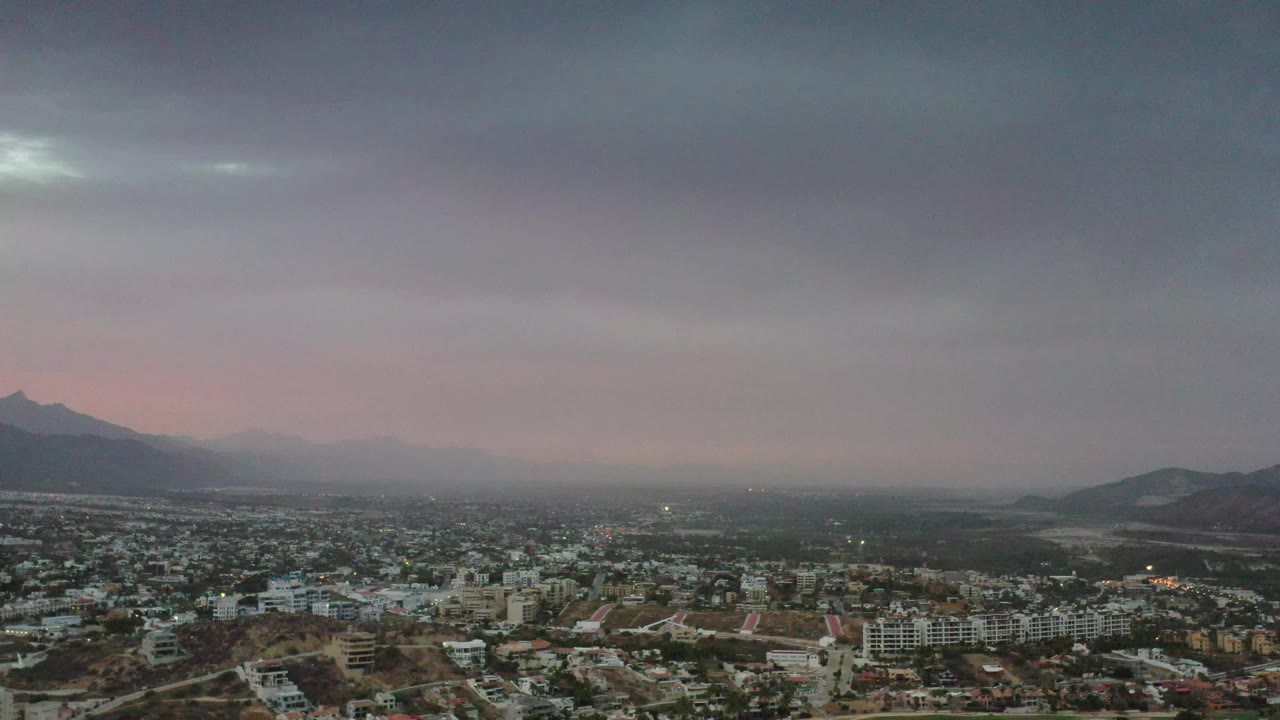 Los Cabos appears from a high vantage, with dense low-rise buildings below, a muted pink band of evening light, distant mountains, and no visible people.