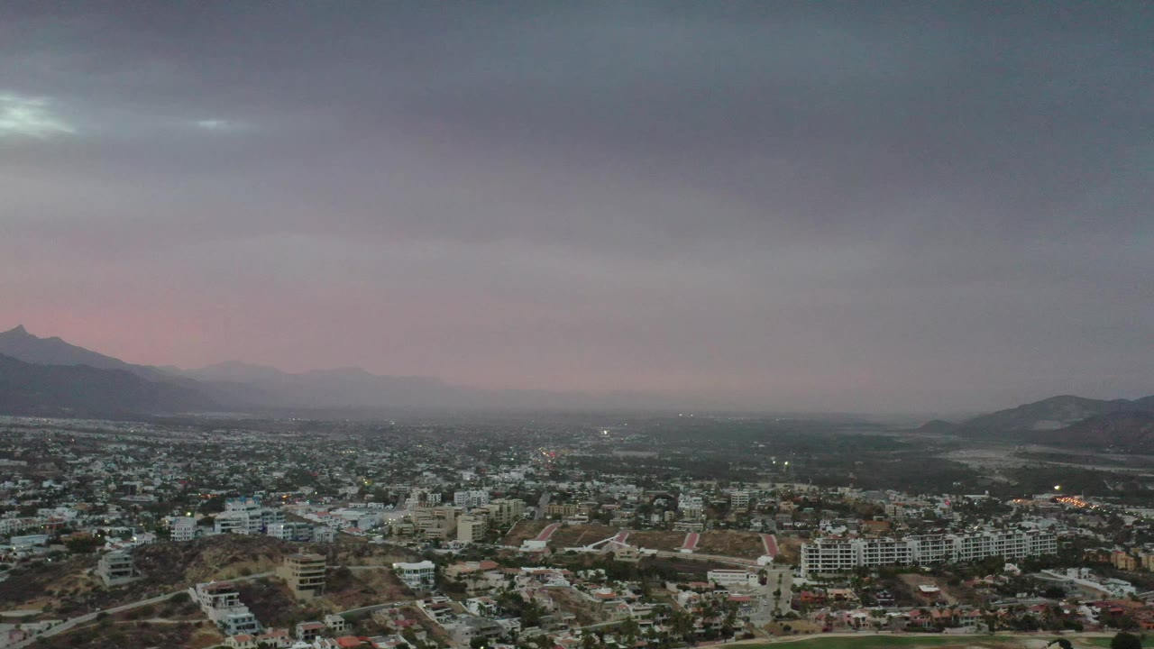 High aerial frame of Los Cabos residential blocks and streets, with darker hills in the midground, a hazy mountain ridge beyond, and no visible people.