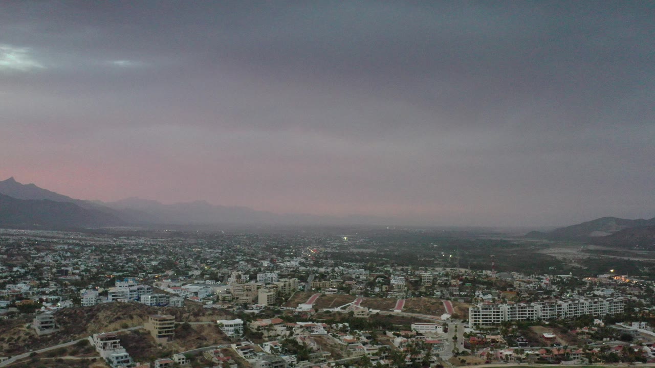 Opening frame of Los Cabos, Mexico, showing a wide urban area, brown hills, distant mountains, pink-gray haze, heavy overcast sky, and no visible people.