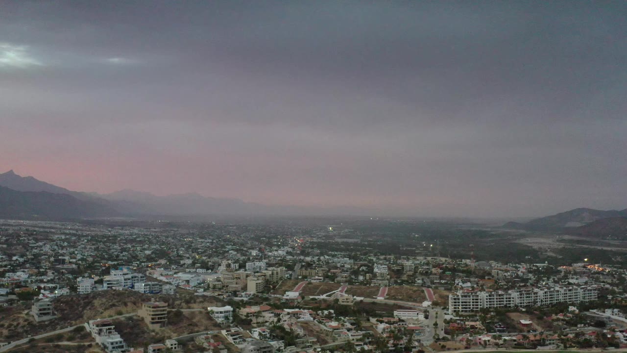 Los Cabos, Mexico, appears under a broad gray cloud layer with low-rise buildings, scattered lights, open desert ground, distant ridges, and no visible people.