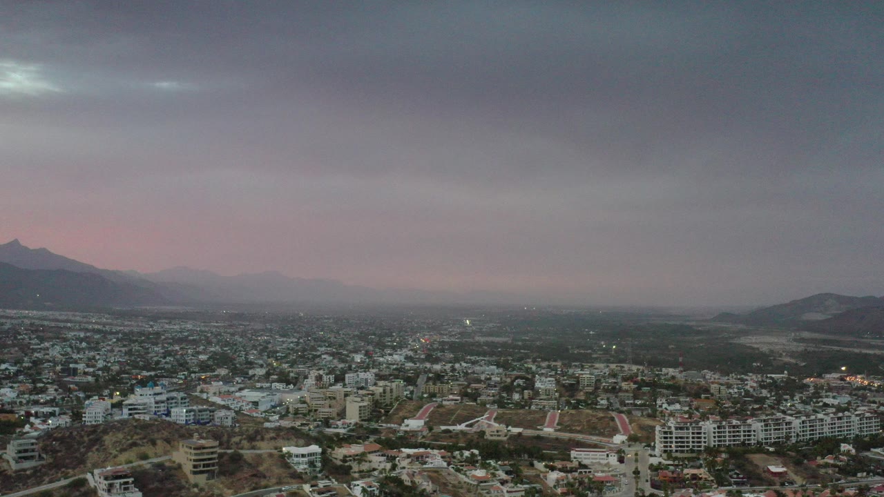 Wide elevated view over Los Cabos at twilight, with low-rise buildings in the foreground, distant mountains on the horizon, a pink sky band, and no visible people.