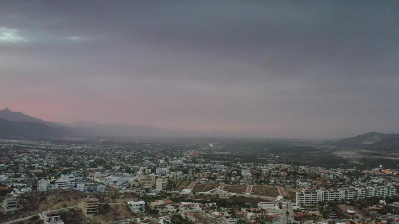Los Cabos city blocks fill the lower frame beneath gray cloud cover, with dark open land, layered mountains, faint evening color, and no visible people.