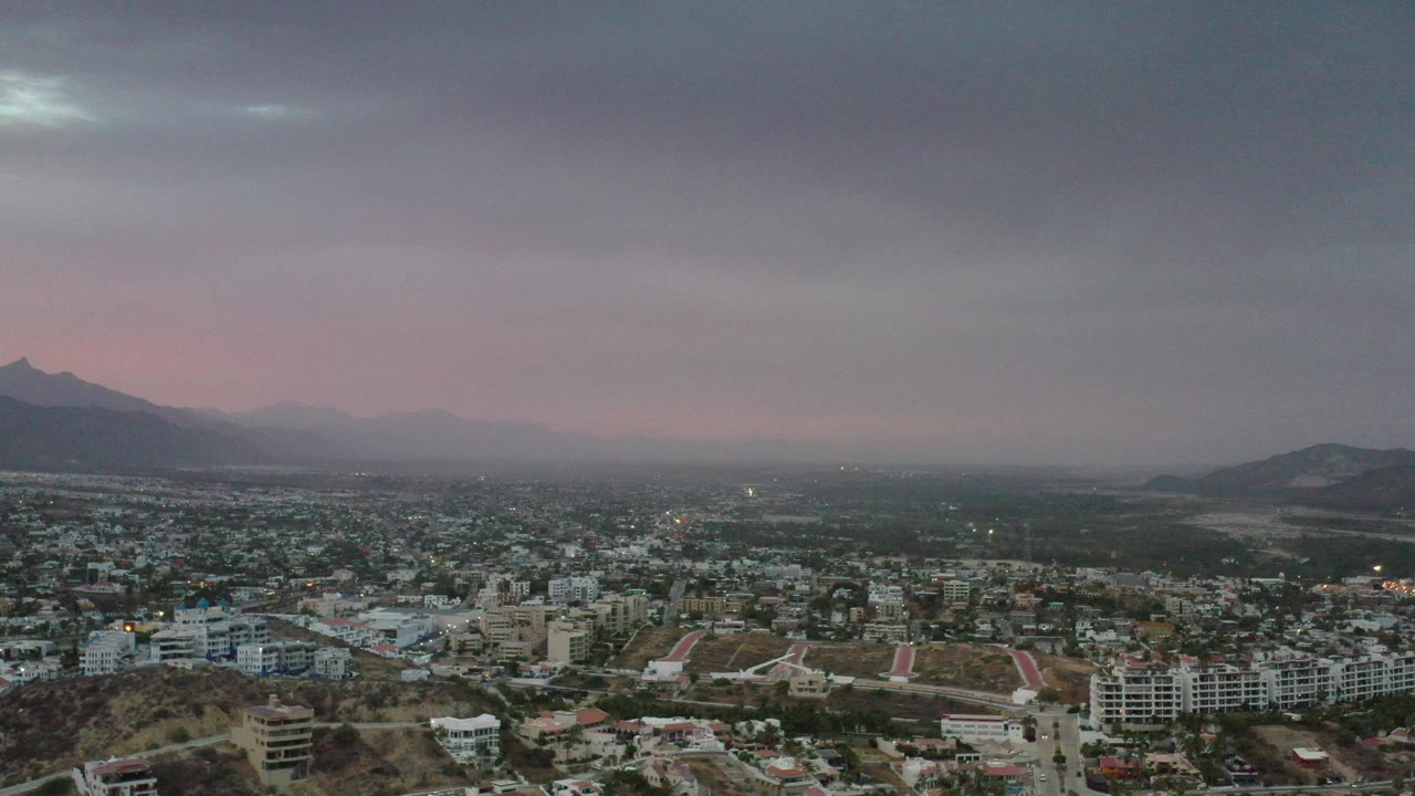 Opening frame of Los Cabos from above, with low hillside foreground, dense city blocks, distant mountains, a pink horizon, and no visible people.