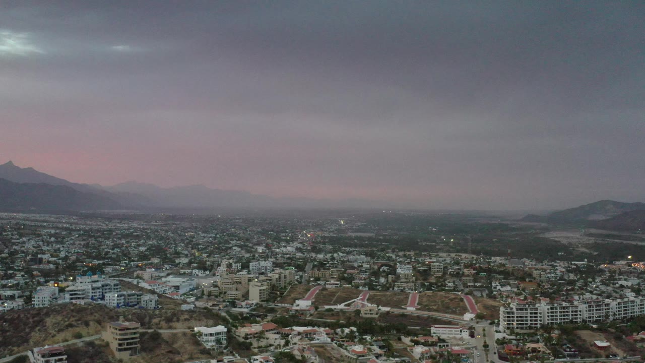 High view over Los Cabos with clustered buildings, hills, and a clouded dawn sky.