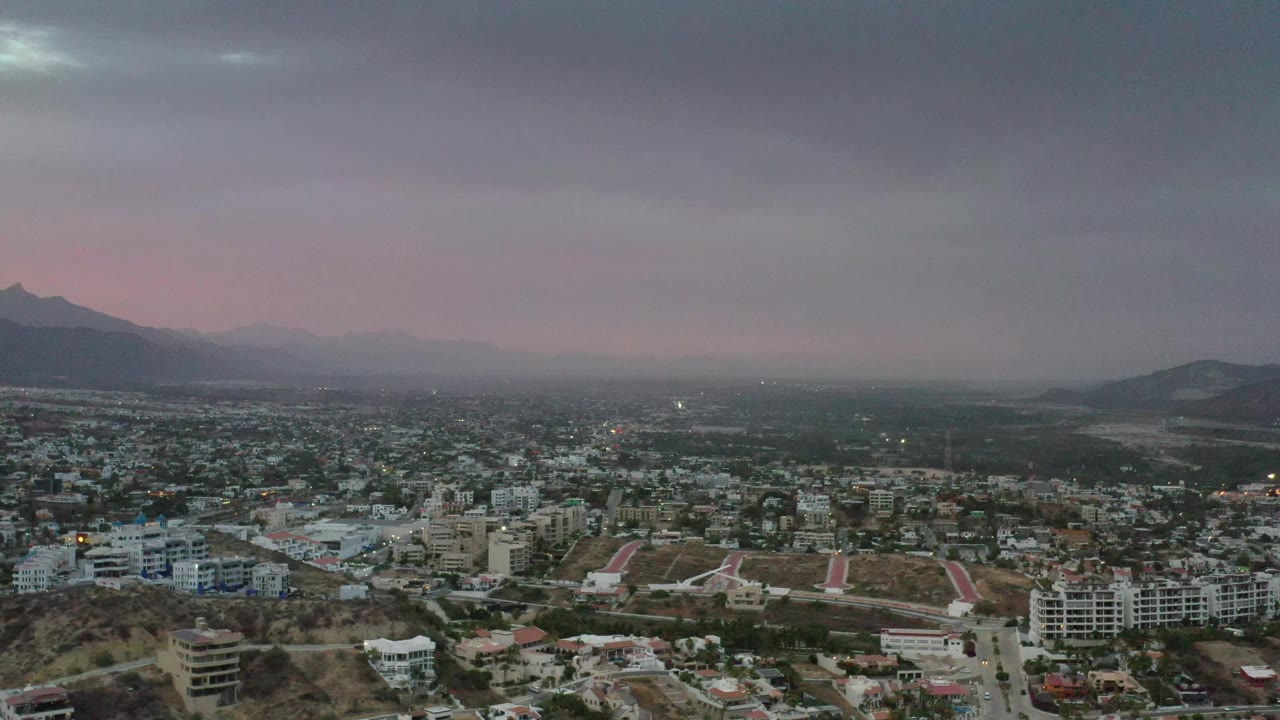 Los Cabos appears from an elevated angle with dense low-rise neighborhoods below, a mountain ridge on the left, layered clouds overhead, and no visible people.