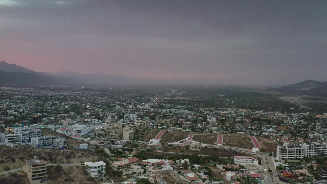 Opening aerial frame of Los Cabos, Mexico, with low-rise buildings, roads, dry hills, distant mountains, muted pink light near the horizon, and no visible people.